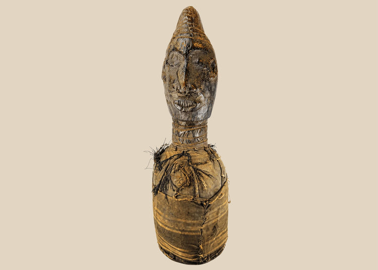 Hand-carved Central or West African wooden figure with elongated facial features, natural fiber torso wrapping, and traces of organic pigments, standing on a white background.