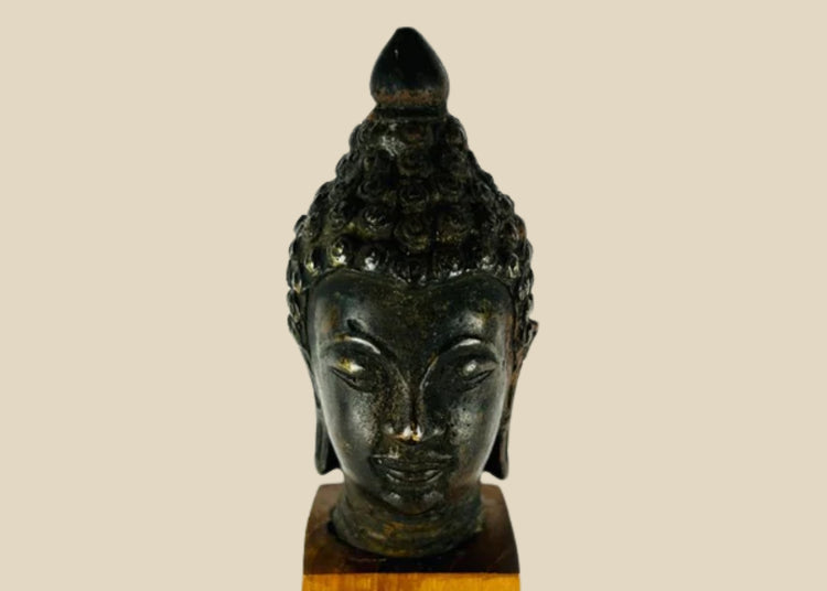 Antique Southeast Asian cast bronze Buddha head, likely from Thailand or Burma, 18th–19th Century CE. Features serene facial expression, closed eyes, gentle smile, ushnisha symbolizing wisdom, and intricately modeled hair curls. Dark natural patina with wooden base, reflecting Theravāda Buddhist devotional artistry and meditation iconography.
