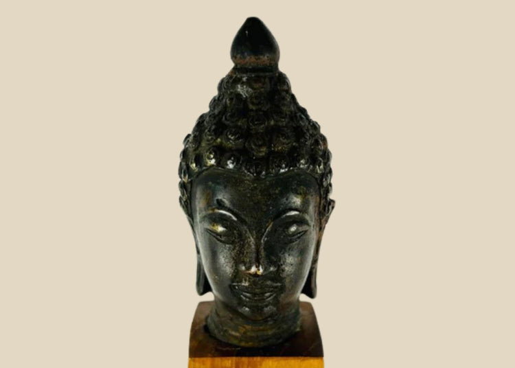 Antique Southeast Asian cast bronze Buddha head, likely from Thailand or Burma, 18th–19th Century CE. Features serene facial expression, closed eyes, gentle smile, ushnisha symbolizing wisdom, and intricately modeled hair curls. Dark natural patina with wooden base, reflecting Theravāda Buddhist devotional artistry and meditation iconography.