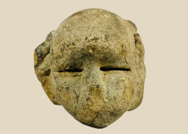 Ancient Mesoamerican stone mask fragment with minimalist facial features and narrow eye slits, carved from a single block of stone and mounted on a black wooden display stand, dating to 500 BCE–500 CE.