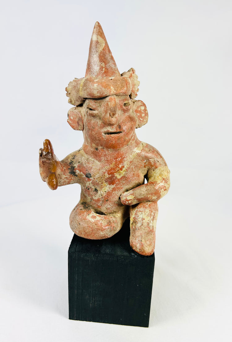Hollow West Mexican Nayarit seated terracotta figure with tall conical headdress, turban band, large earspools, and raised right hand holding an attribute, featuring red and cream slip remnants, mineral accretions, and mounted on a modern wood display block, ca. 100 BCE–300 CE