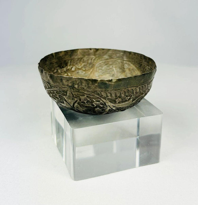 Angled view of Seljuk Persian silver unguent bowl from 12th–13th century CE, featuring intricate engraved floral and geometric motifs with aged silver patina, displayed on a clear acrylic base under studio lighting.