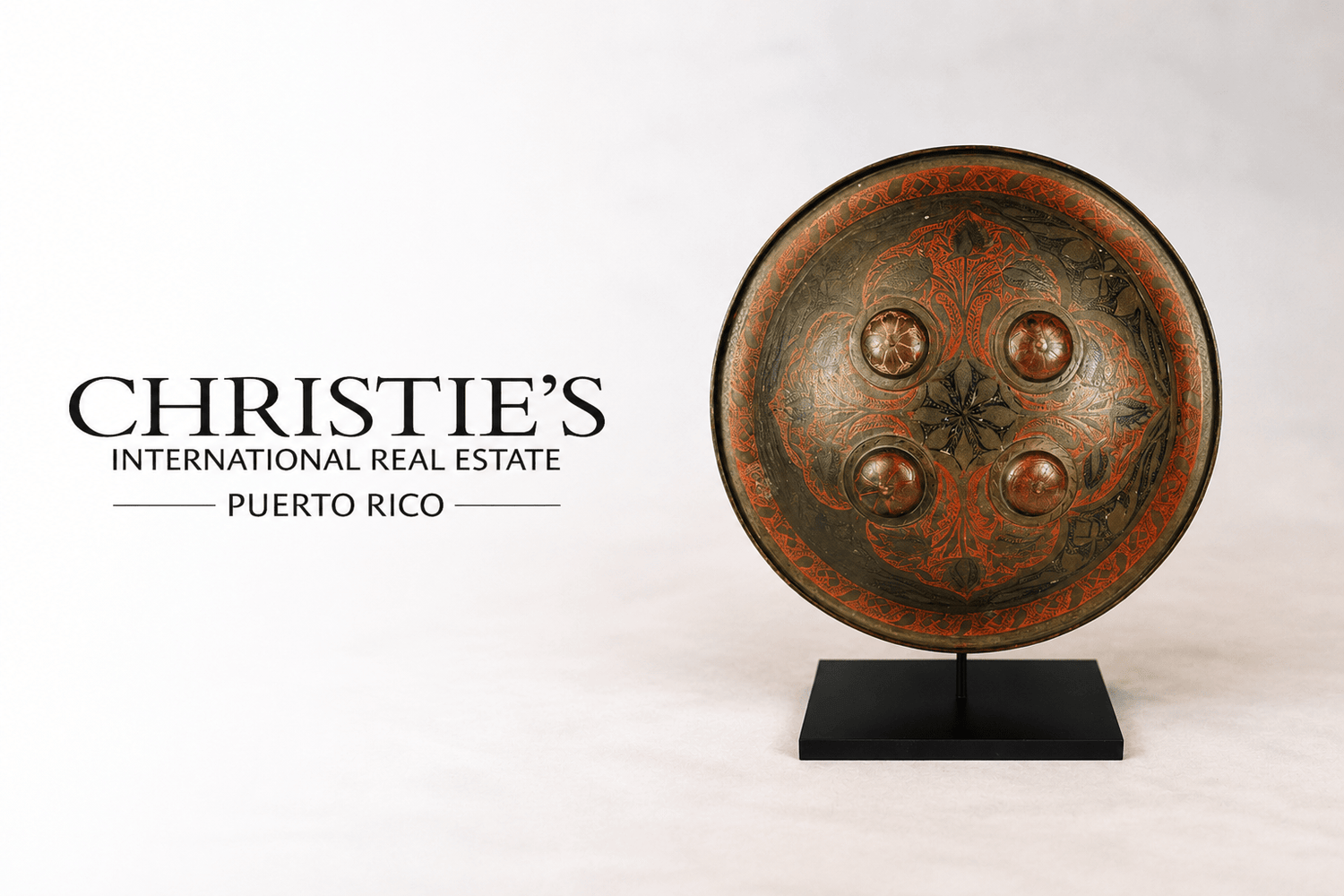 Ancient decorative shield displayed beside Christie’s International Real Estate Puerto Rico logo on a soft neutral studio background, featuring intricate red and black ornamental detailing on a museum-style stand.