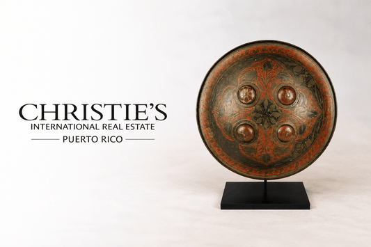 Ancient decorative shield displayed beside Christie’s International Real Estate Puerto Rico logo on a soft neutral studio background, featuring intricate red and black ornamental detailing on a museum-style stand.