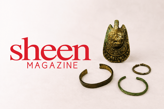 Ancient bronze and copper artifacts arranged on a soft neutral studio background beside the red Sheen Magazine logo, featuring a weathered patinated amulet and three ancient bracelets with visible age and oxidation.