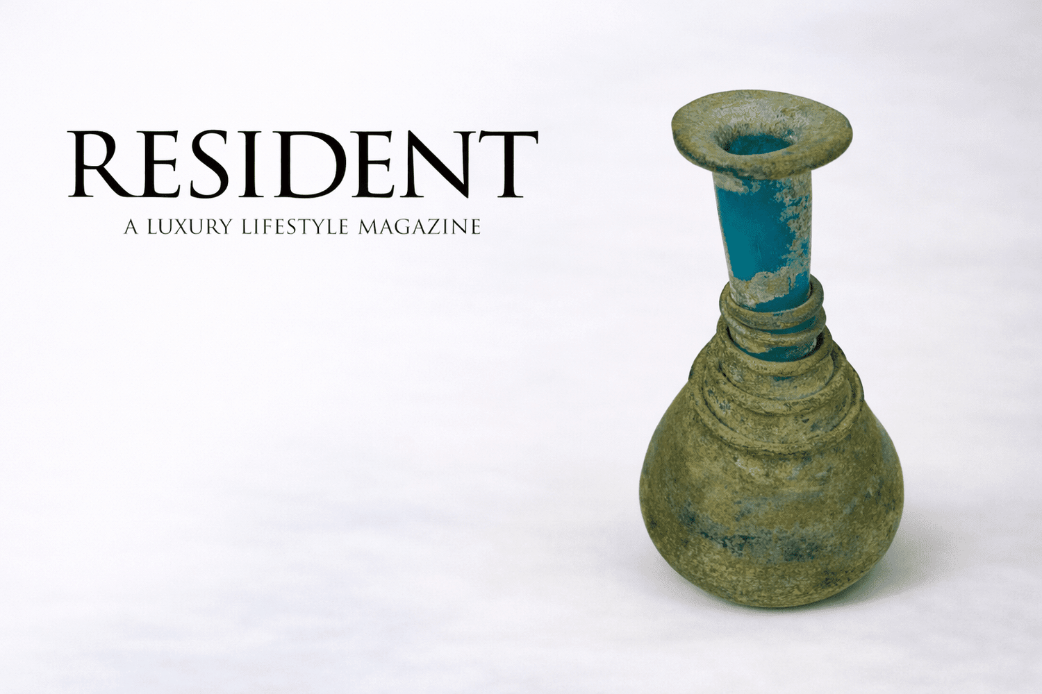 Resident luxury lifestyle magazine logo displayed beside an ancient Roman turquoise glass perfume bottle with spiral rings from the 1st–3rd century CE against a neutral studio background.