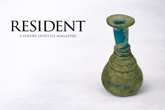 Resident luxury lifestyle magazine logo displayed beside an ancient Roman turquoise glass perfume bottle with spiral rings from the 1st–3rd century CE against a neutral studio background.