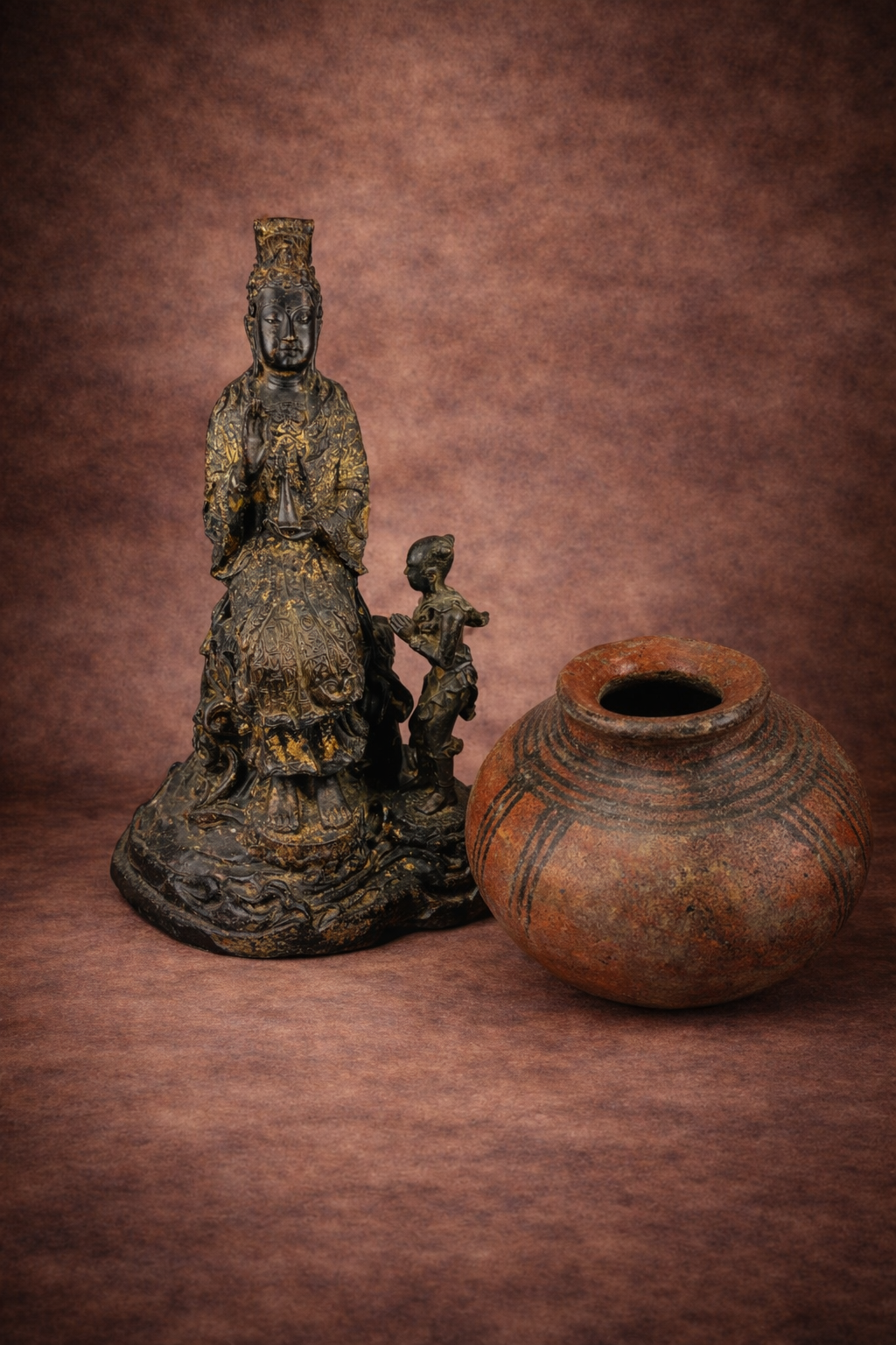 20th Century antique gilt bronze Bodhisattva sculpture with attendant figure displayed beside a West African terracotta pot featuring painted geometric decoration, photographed together against a warm neutral studio background.
