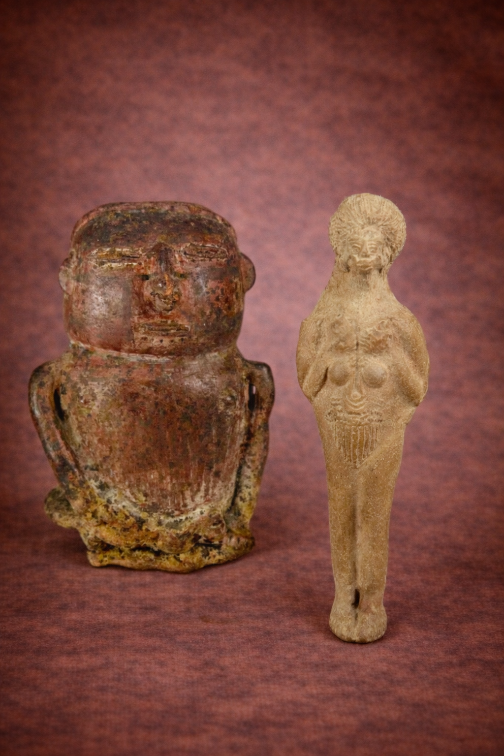 Pre-Columbian terracotta effigy vessel and ancient Roman terracotta fertility idol displayed side by side on a warm reddish studio background, highlighting surface wear, earthen tones, and archaeological patina.