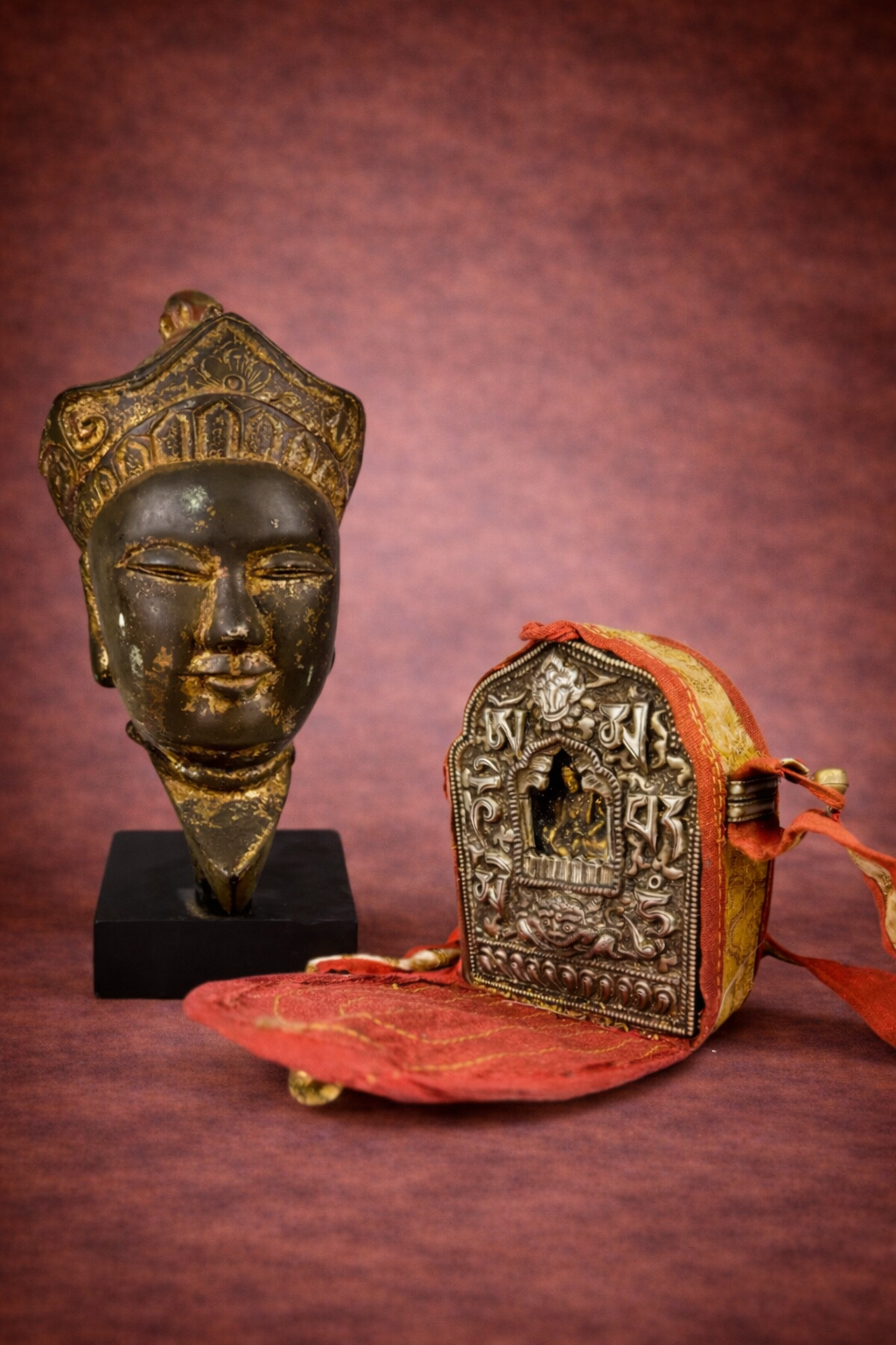 Tibetan Buddha head sculpture displayed beside an antique Buddhist gau reliquary shrine on a neutral background.