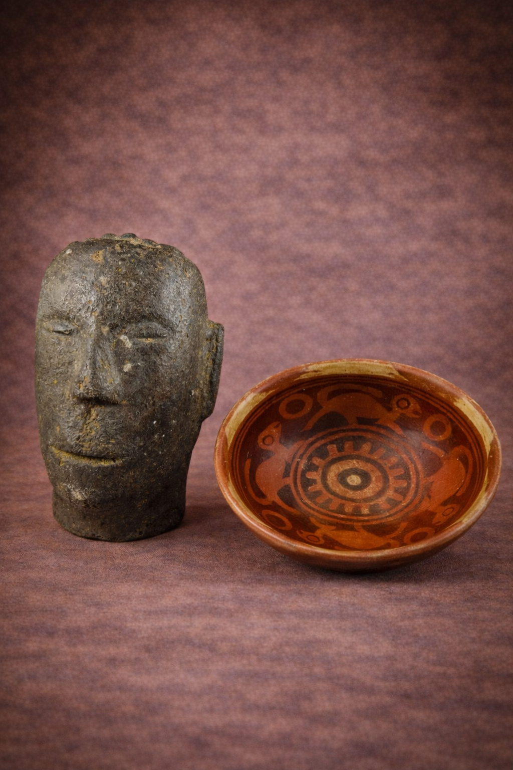 Polynesian volcanic stone ancestor head displayed beside a Pre-Columbian Nariño red-slip pottery bowl on a warm brown studio background.