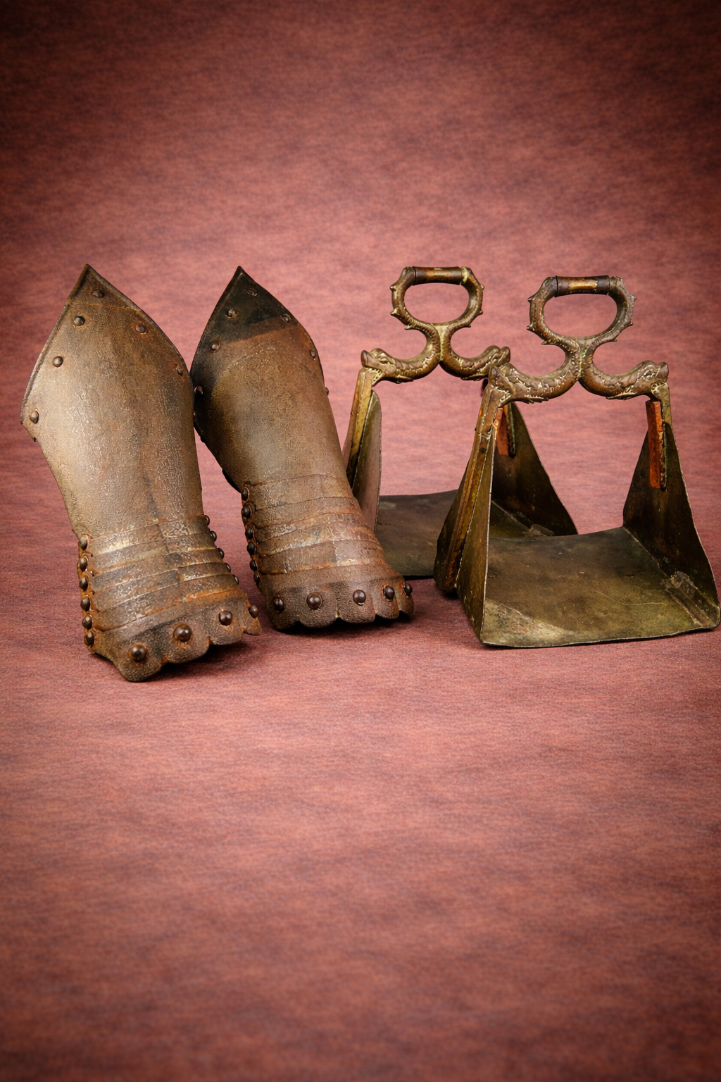Pair of Gothic-style steel gauntlets displayed beside ornate brass equestrian stirrups with serpent motifs on a warm brown studio background.