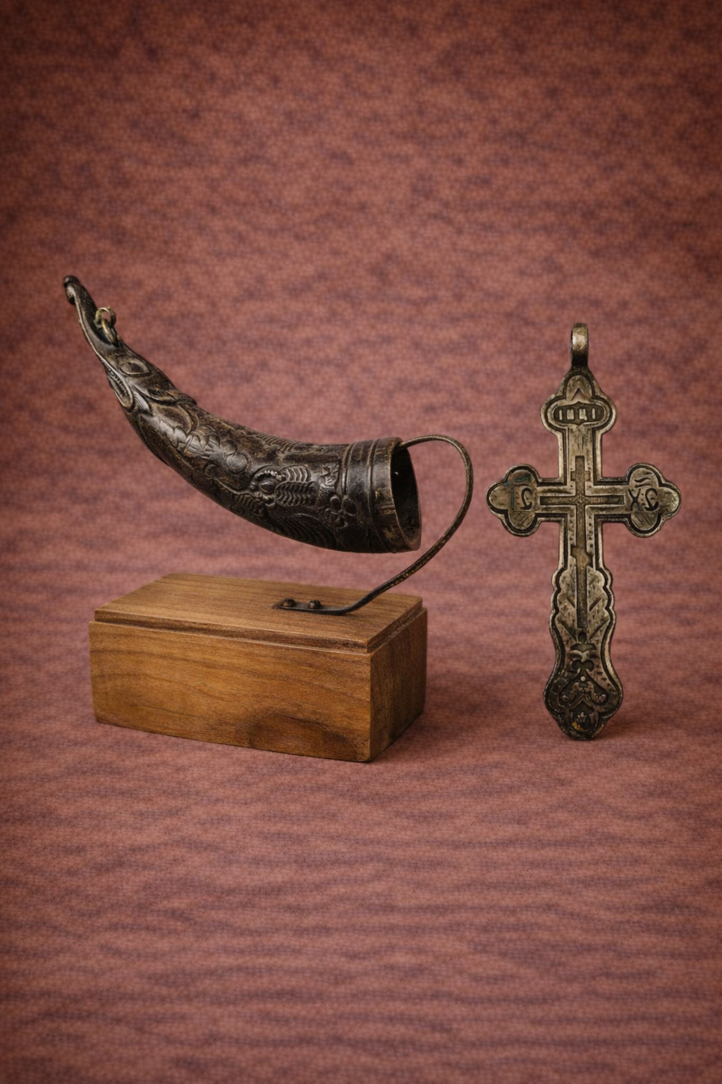 Tibetan ritual horn displayed on a wooden stand beside an antique Russian Orthodox silver cross, both photographed against a warm textured studio backdrop.