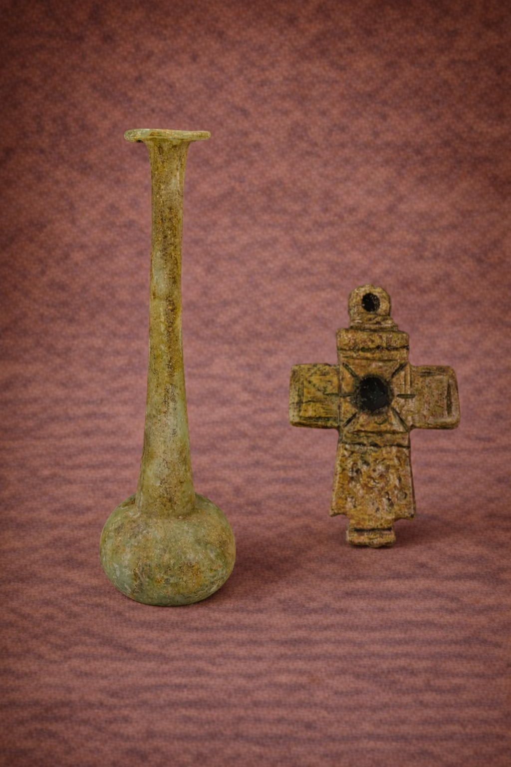 Ancient Roman glass perfume bottle displayed beside a Byzantine pectoral cross pendant, both photographed against a warm textured studio background showing age wear and surface patina.