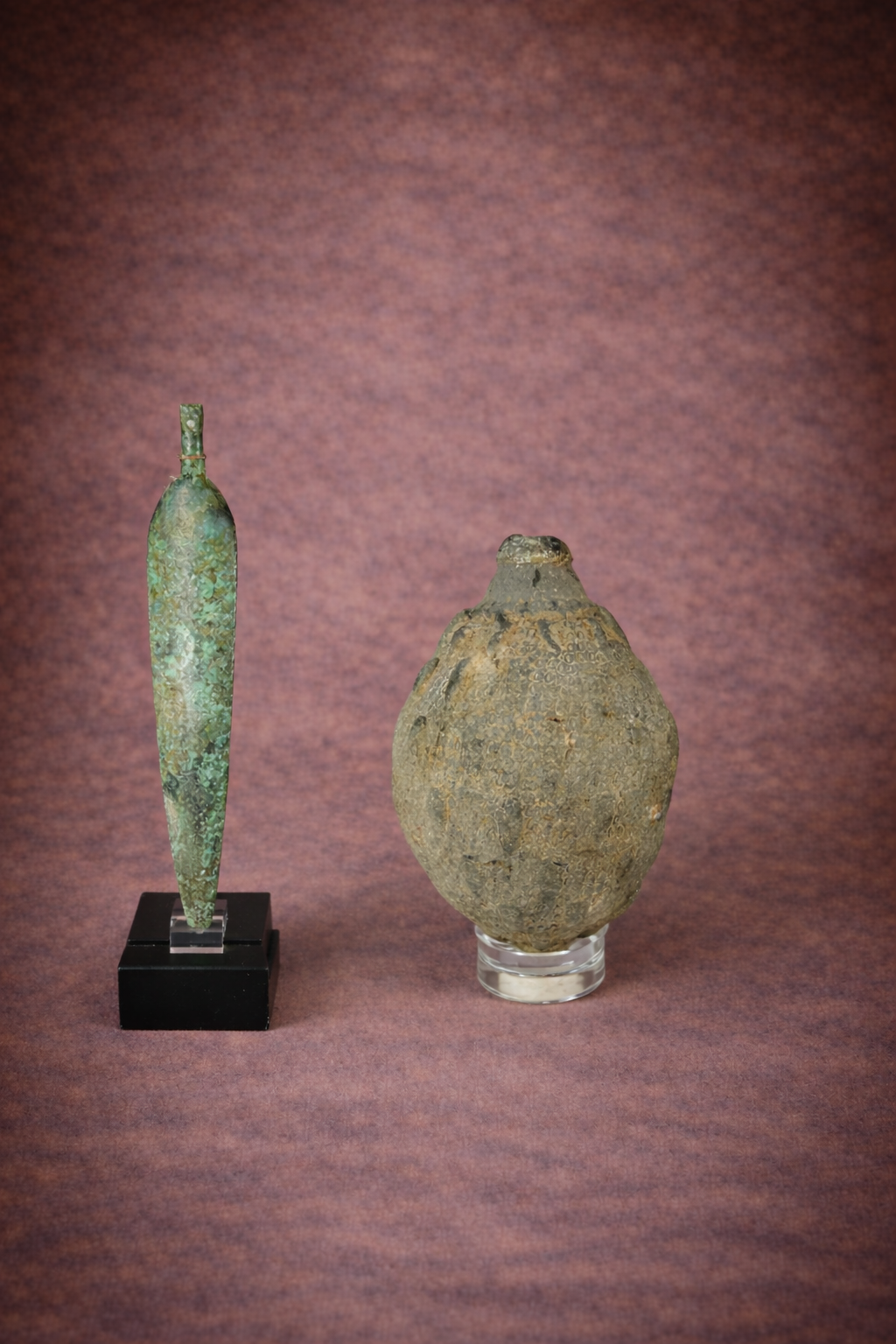 Ancient bronze dagger with green patina displayed on a stand beside a weathered ceramic hand grenade, both photographed against a textured brown studio backdrop.