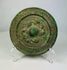 Ancient Han Dynasty bronze mirror with green patina, circular motifs, and raised central knob displayed on a clear stand.