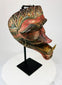 Hand-carved Javanese Cakil mask with aged painted details, displayed at an angle on a black stand.
