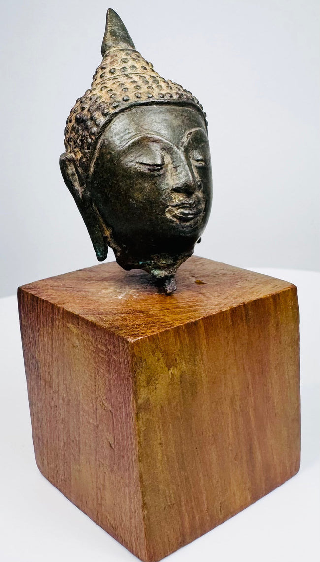 Thai bronze Buddha head with downcast eyes, a gentle smile, and dark green-brown patina, displayed on a square wooden base.