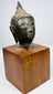 Thai bronze Buddha head with downcast eyes, a gentle smile, and dark green-brown patina, displayed on a square wooden base.