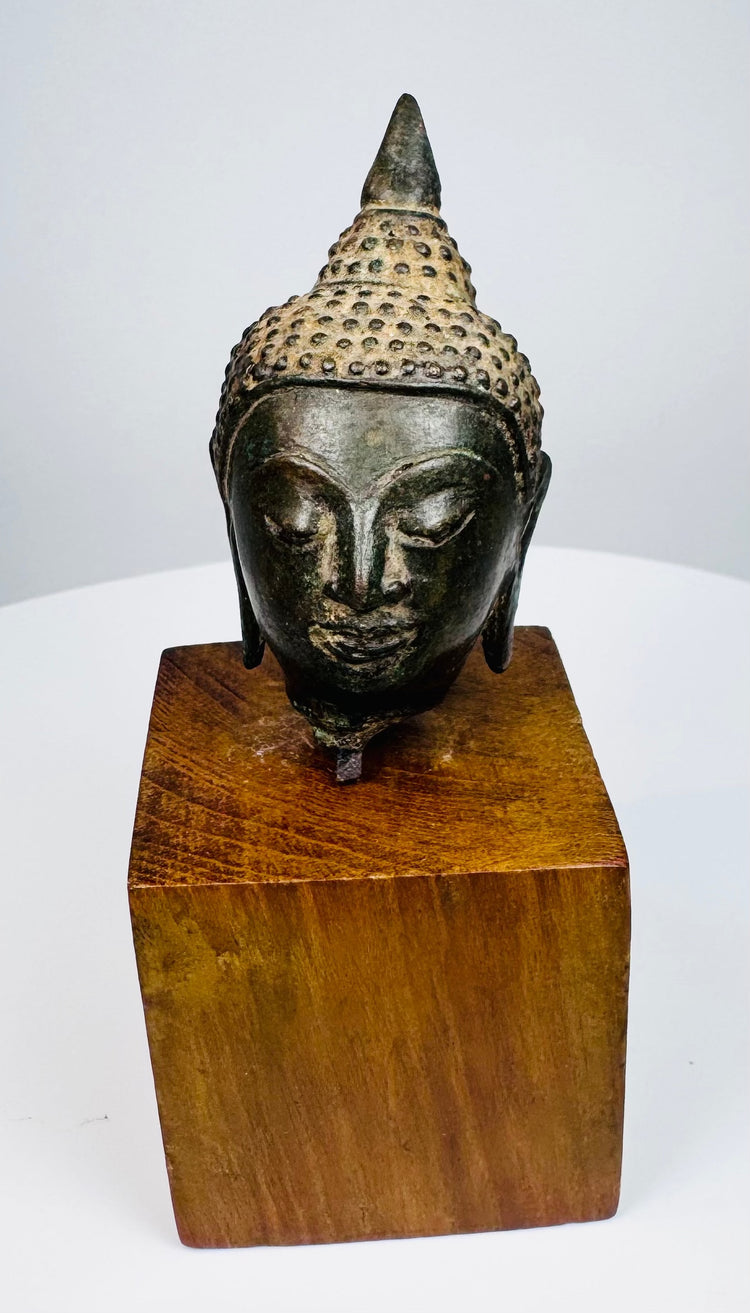 Bronze Buddha head with a calm expression, tightly patterned curls, and dark patina, mounted on a rectangular wooden base.