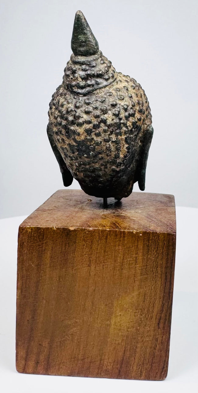 Thai bronze Buddha head with pale surface wear contrasting against dark curls across the rounded head, set on a wooden base.