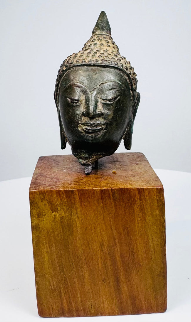 Thai bronze Buddha head displaying arched brows, softly curved lips, and elongated earlobes above a warm brown wooden block.