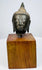 Thai bronze Buddha head displaying arched brows, softly curved lips, and elongated earlobes above a warm brown wooden block.