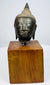 Thai bronze Buddha head displaying arched brows, softly curved lips, and elongated earlobes above a warm brown wooden block.