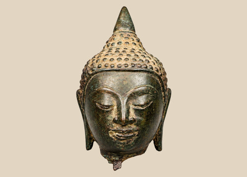 Thai bronze Buddha head with half-closed eyes, elongated earlobes, and a pointed topknot against a beige background.
