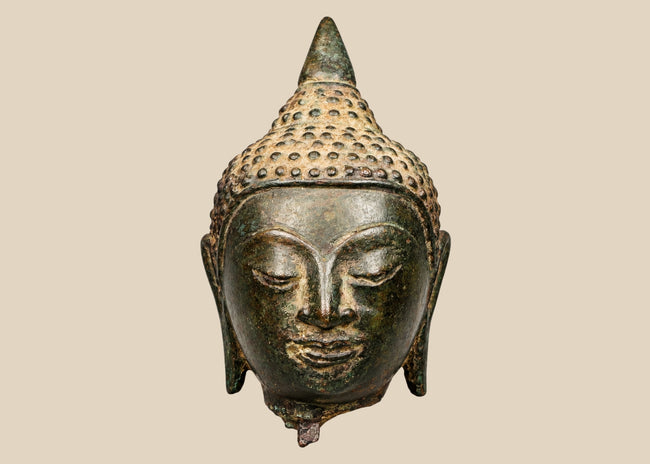 Thai bronze Buddha head with half-closed eyes, elongated earlobes, and a pointed topknot against a beige background.