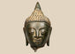 Thai bronze Buddha head with half-closed eyes, elongated earlobes, and a pointed topknot against a beige background.