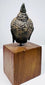 Bronze Buddha head showing dense rows of curls, mottled surface patina, and a pointed topknot above its wooden mount.
