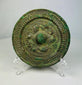 Ancient Han Dynasty bronze mirror with green patina, circular motifs, and raised central knob displayed on a clear stand.