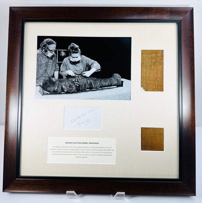 Dark wood frame displaying two attributed Manchester Mummy 1770 linen fragments beside a photograph of the 1975 investigation.