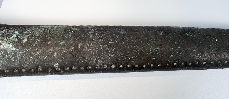 Metal sword sheath with red and dark cabochon inserts and green surface corrosion.
