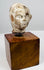Ancient terracotta head with a youthful expression, gently curved lips, and worn facial contours, displayed on a wooden base.