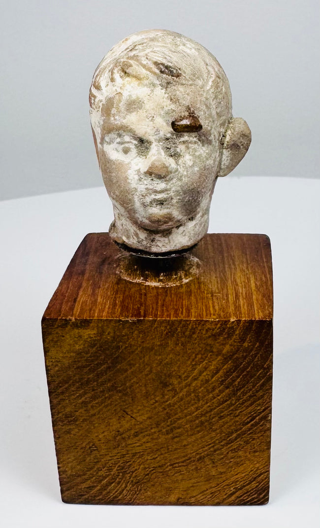 Terracotta head fragment with sculpted eyes, softly curved lips, and worn hair detailing, mounted on a warm brown wooden block.