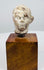 Ancient terracotta head displaying rounded cheeks, defined brows, and a prominent ear above a square wooden display base.
