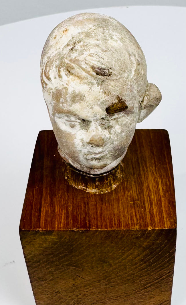 Ancient terracotta head showing sculpted hair, recessed eyes, and a small mouth above its warm brown wooden display mount.