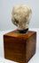 Ancient terracotta head with a rounded crown, weathered surface, and uneven neck edge above a brown wooden display base.