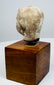 Ancient terracotta head with a rounded crown, weathered surface, and uneven neck edge above a brown wooden display base.