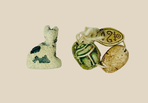 Ancient Egyptian faience cat amulet with traces of blue-green glaze, shown alongside three scarabs in varied materials, on a white background.