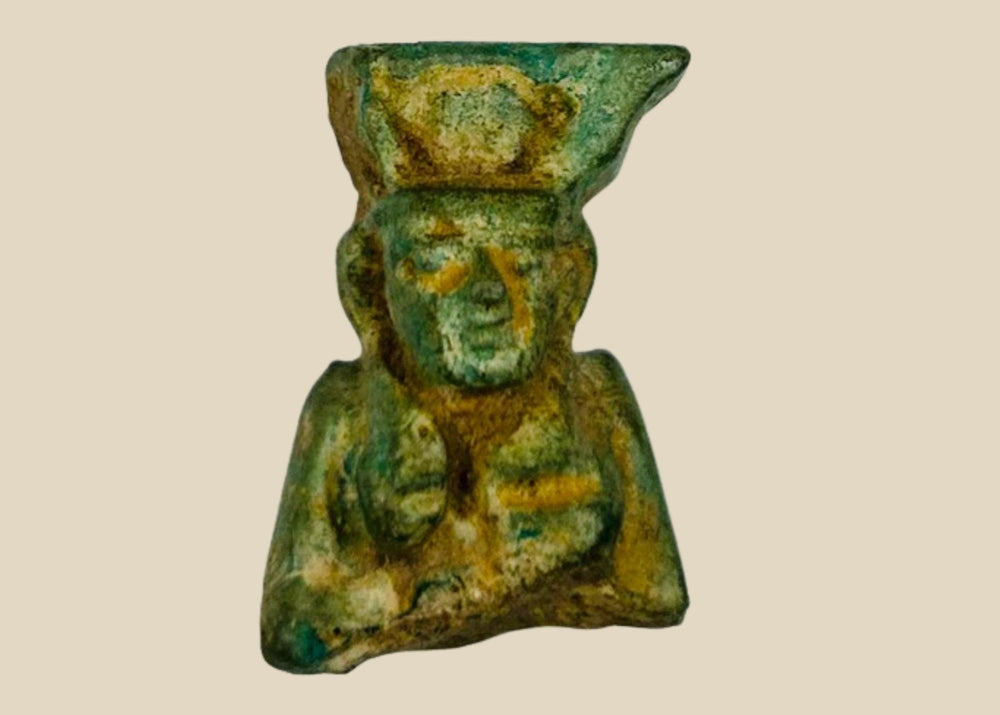 Miniature pale blue-green faience amulet of lion-headed goddess Sekhmet, front-facing with tripartite wig and crown traces; suspension hole on reverse; mounted on black wood stand; Egypt, Third Intermediate Period, circa 1070–664 BCE.
