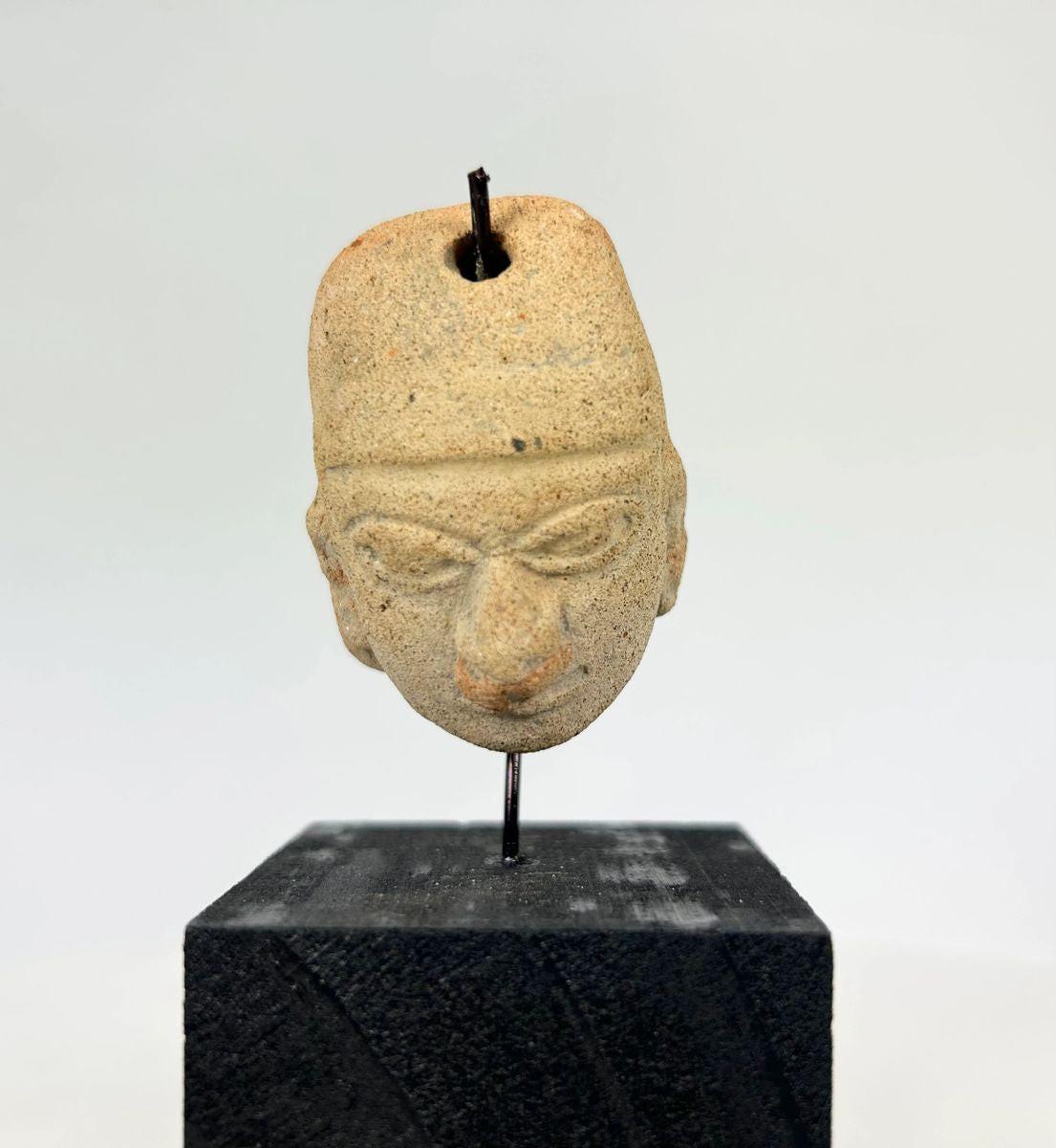 Terracotta pendant head with angular headdress, incised almond-shaped eyes, furrowed brow, and pronounced nose and lips; drilled suspension hole through top; mounted on a museum-style stand; West Mexico (Colima/Nayarit) or Veracruz, circa 300 BCE–300 CE.
