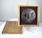 Boxed Japanese tsuba showing mixed-metal relief decoration, central tang opening, and purple fitted lining