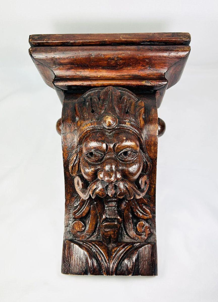 Antique 19th-century European wooden corbel, hand-carved with a grotesque crowned face featuring a prominent mustache and ornamental details. Dark natural patina, Gothic-inspired design, and architectural functionality. Serves as both structural support and decorative element, reflecting historic woodcarving traditions and Gothic revival aesthetics.