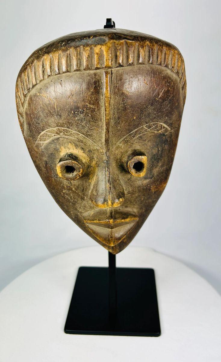 Antique Baule tribal mask from Ivory Coast, early 20th century, hand-carved wood with natural patina, oval face, almond-shaped eyes, and stylized features displayed on a stand.