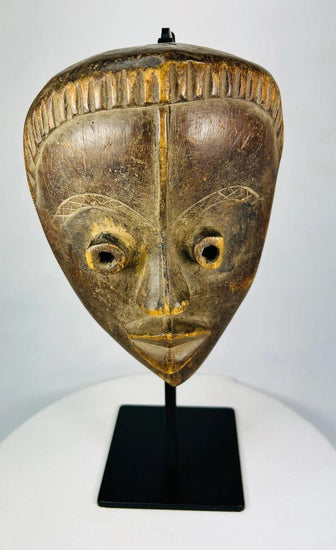 Antique Baule tribal mask from Ivory Coast, early 20th century, hand-carved wood with natural patina, oval face, almond-shaped eyes, and stylized features displayed on a stand.