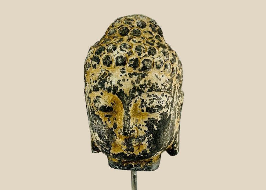 Antique carved stone Buddha head from Southeast Asia, likely Thailand or Cambodia, 18th–19th Century. Features serene meditative expression, curled hair, and traces of original pigments. Mounted on a marble base, reflecting Buddhist artistry, spiritual symbolism, and historical craftsmanship of the region.