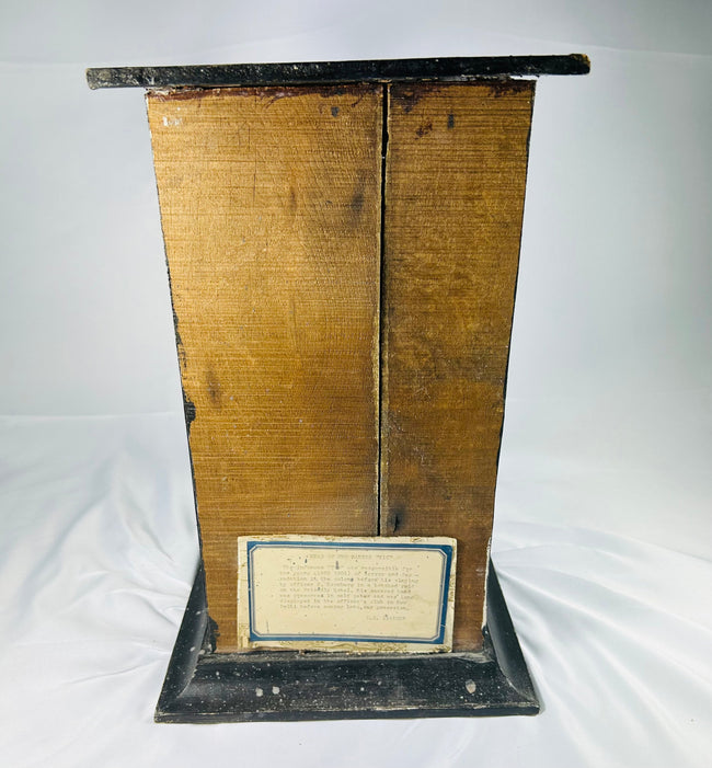 Rear view of 19th-century mummified head display case showing aged wooden backing and brass plaque with historical inscription on a white background.